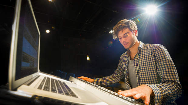 Man playing a digital piano with a laptop in front of him under stage lights.
