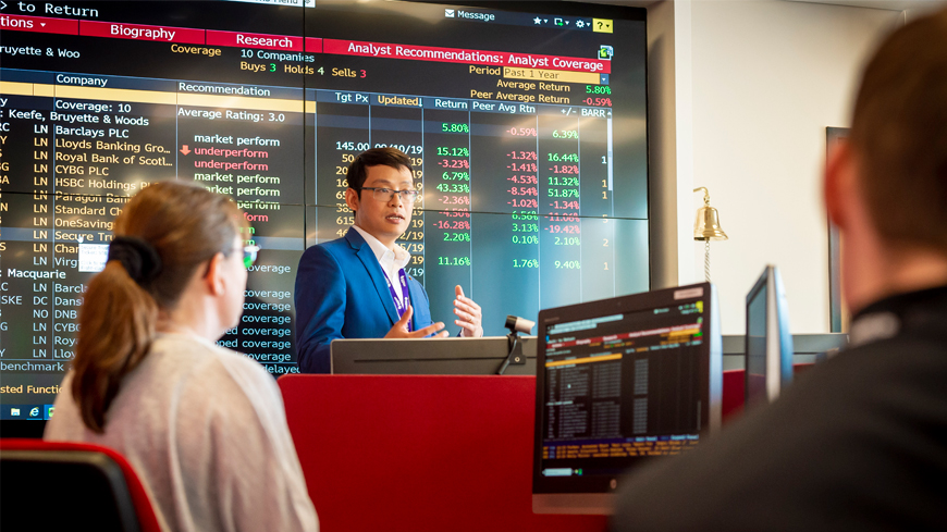 An academic speaks to a class in front of a large stock trading board