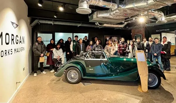 A large group of students standing around a dark green vintage car next to a Morgan sign