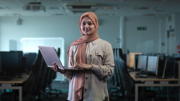 Student holding a laptop with a computer lab in background