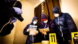 Investigators in protective clothing and masks examine evidence marked with numbered signs indoors.
