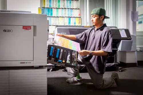 Person kneeling and inspecting a large color print in front of a commercial printer in an office or print shop.