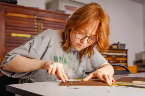 Person with red hair using a ruler and utility knife to cut material on a worktable in a workshop or studio setting.