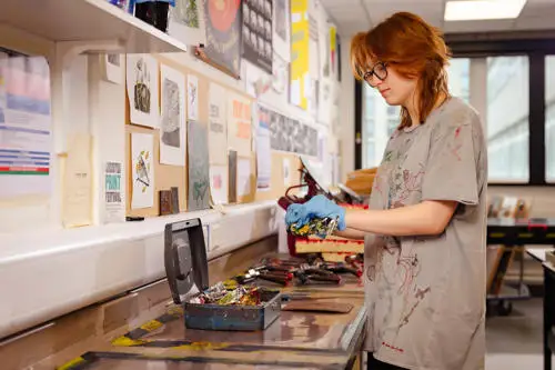 Person wearing gloves works with art supplies at a cluttered studio table, with various tools and artworks visible on the wall.