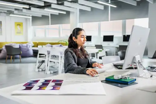 Woman working at a computer in a bright, modern office with charts and documents on her desk.