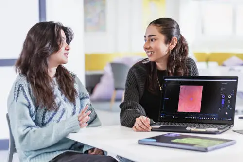 Two women sitting at a table talking, with one showing a document on a laptop screen.
