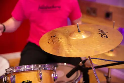 Person playing a drum set, focusing on a cymbal and the drummer's arm in a bright pink shirt.