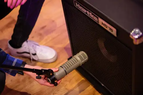 A person positions a microphone in front of a guitar amplifier on a wooden floor.