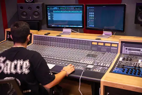 Person wearing headphones operates an audio mixing console in front of computer monitors displaying audio editing software.