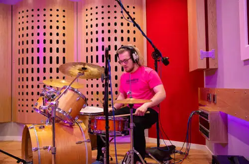 Person playing a drum set in a recording studio, wearing headphones and a pink shirt, with a microphone overhead.