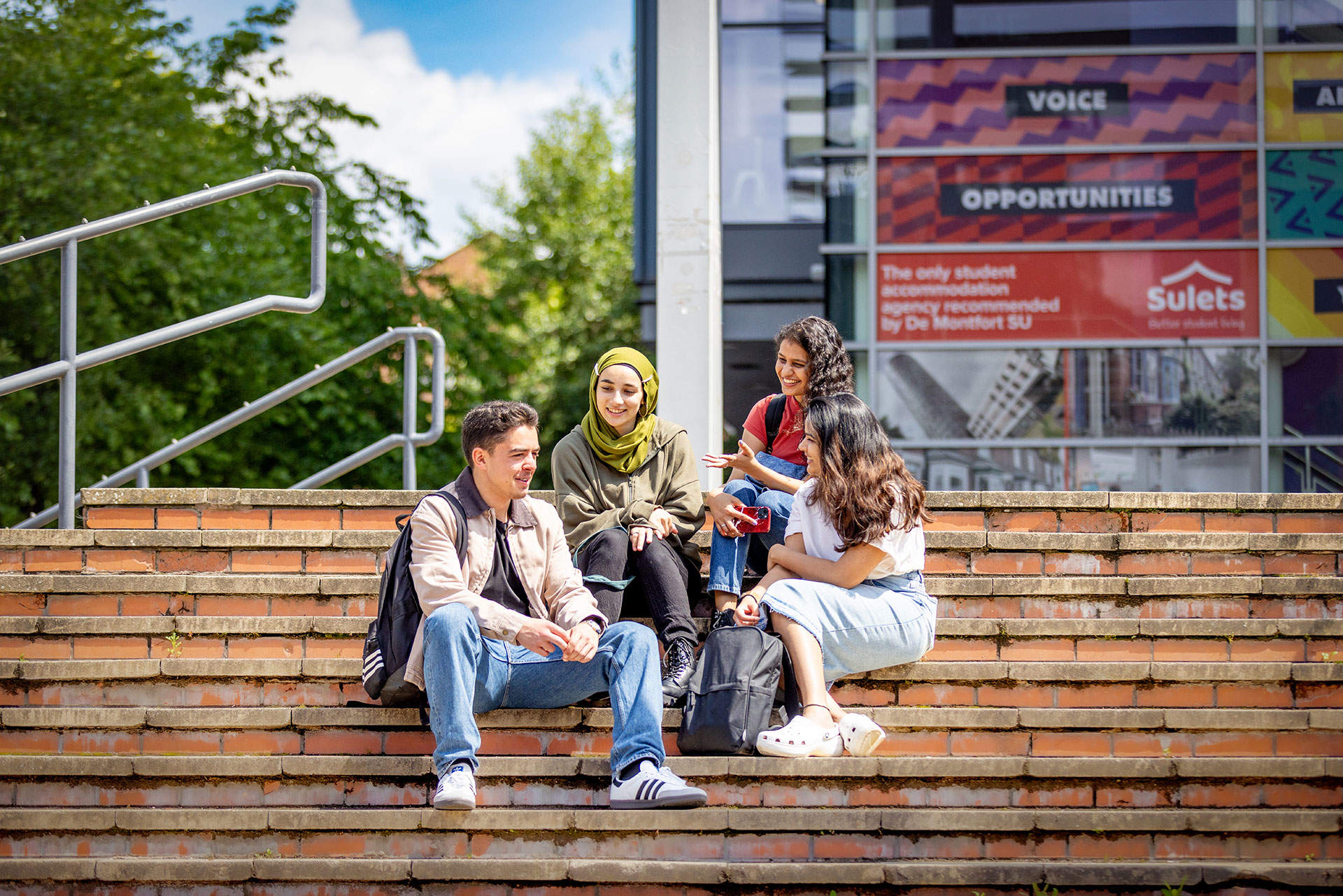 Four young people sit on outdoor steps with backpacks, talking and smiling, with colorful posters in the background.