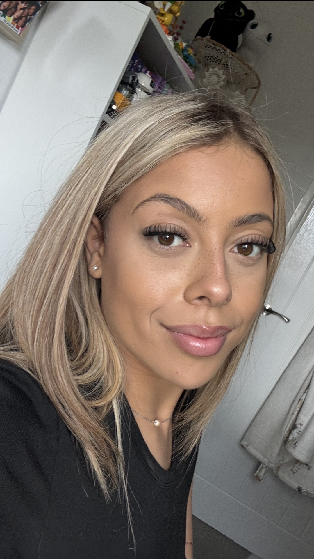 Woman with straight blonde hair and winged eyeliner, wearing a black top, looking at the camera indoors.