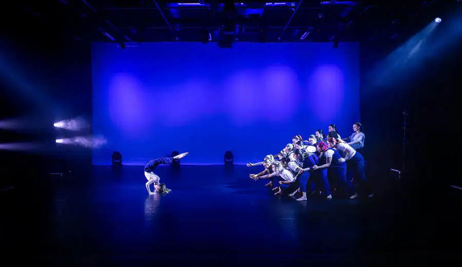 One dancer on the left side of a blue stage and a group of dancers on the right reaching for them