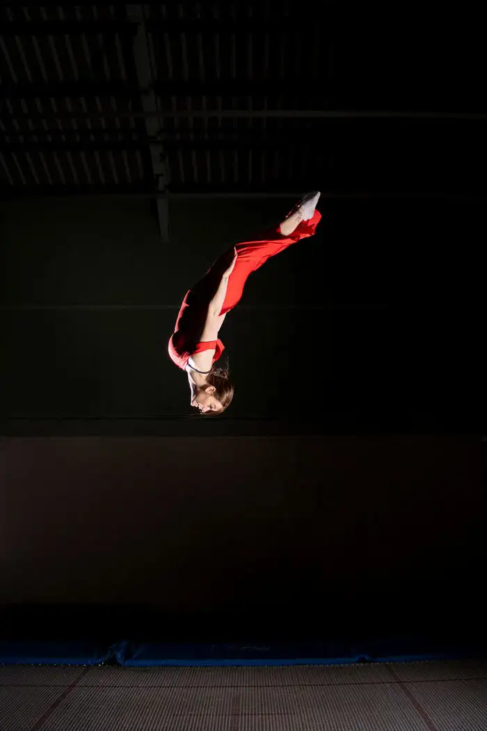 An action shot of a female figure in a red outfit upside down in mid air in a dark gymnasium - by Elianne Grant