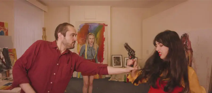 Two people in a room, one holding out a gun to the other, with colorful artwork visible in the background.