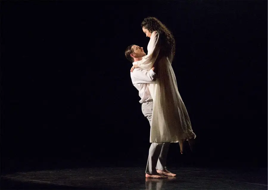 A man lifts a woman in a dance pose on a dark stage, both wearing light-colored clothing.
