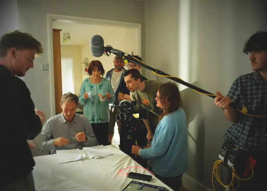 Film crew recording a scene indoors, with a boom microphone operator and several people gathered around a table.