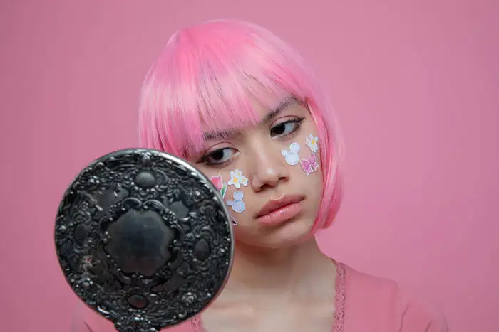 A woman in a pink wig and pink clothing with stickers on her cheeks looking into a black hand mirror on a pink background - by Hanna Milton