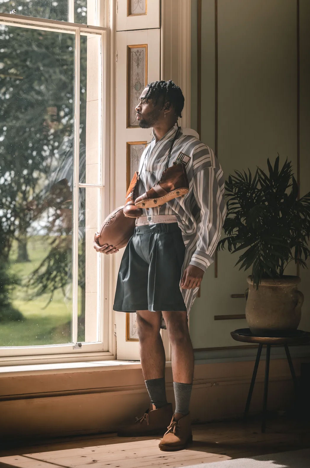 A man holding a rugby ball standing in front of a window; neutral colour palette.