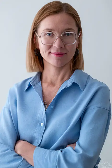 Woman with shoulder-length light brown hair, glasses, and a blue button-down shirt standing with arms crossed.