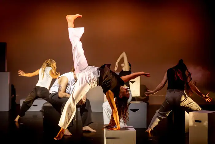 Group of dancers performing dynamic movements on stage with dramatic lighting and props.