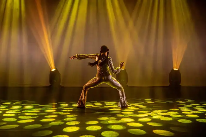 Dancer performing on stage with dramatic yellow lighting and spotlights creating patterns on the floor.