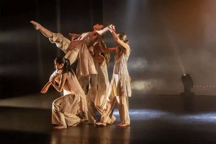 Group of dancers in beige costumes performing a modern dance routine on stage with dramatic lighting.