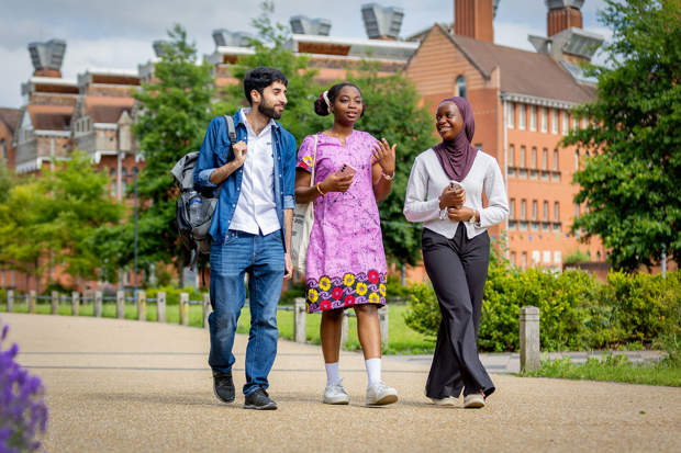 Three young adults walk together outdoors on a campus, smiling and talking, with buildings and greenery in the background.