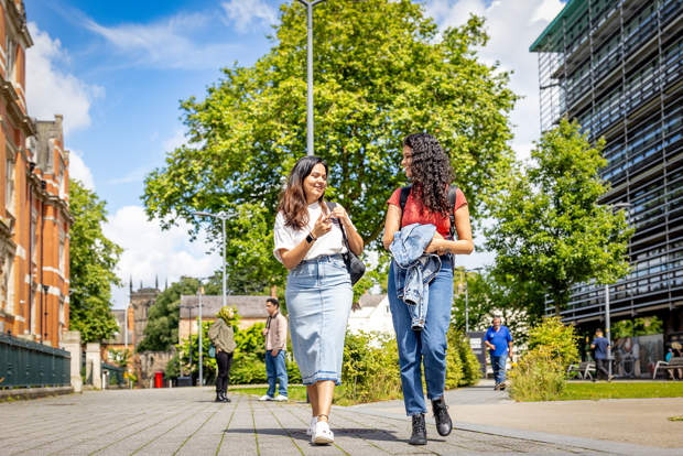 Two young women walking and talking on a sunny campus path, with buildings and trees in the background.