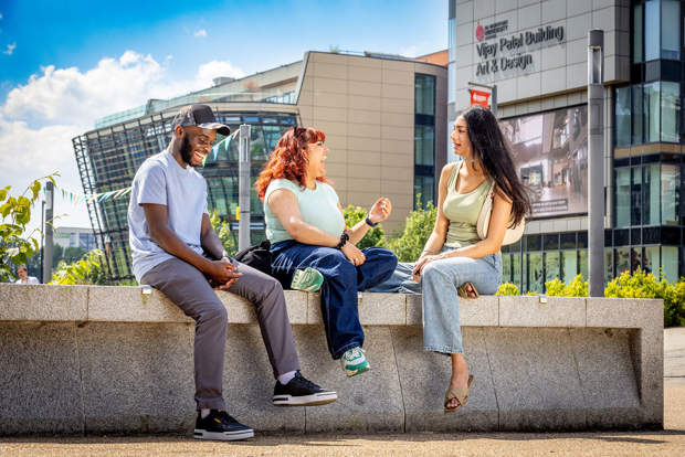 Three young adults sit and talk on a concrete bench outside a modern university building on a sunny day.
