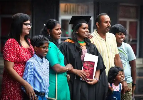 A graduate in a cap and gown holds a diploma, surrounded by smiling family members posing for a photo.