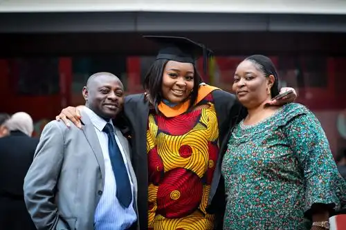 Graduate in cap and gown smiling with two adults standing together and posing for a celebratory photo.