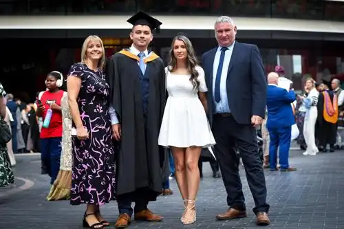 Four people pose together at a graduation ceremony, with one wearing a cap and gown and others dressed formally.