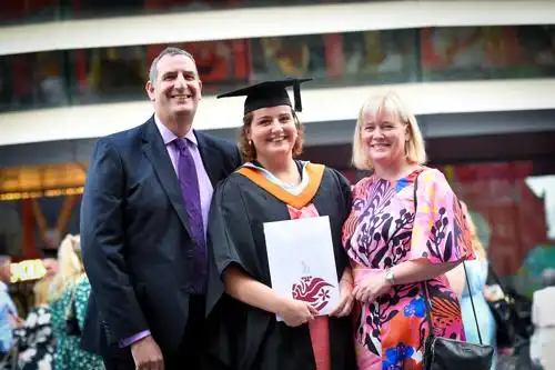 Graduate in cap and gown holding diploma, standing between two smiling adults