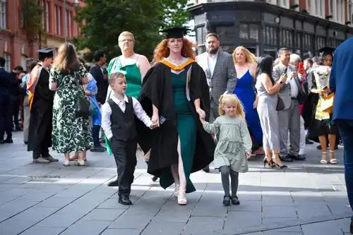 A graduate in cap and gown walks with two children and a group of people on a city street.