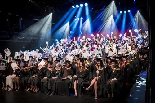 Large audience seated in a theater under bright stage lights, watching an event or performance.