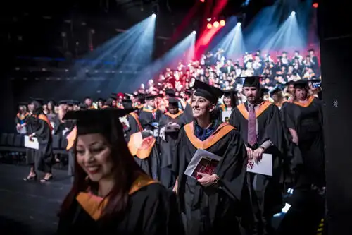 Graduates in caps and gowns stand and smile during a commencement ceremony with stage lights overhead.