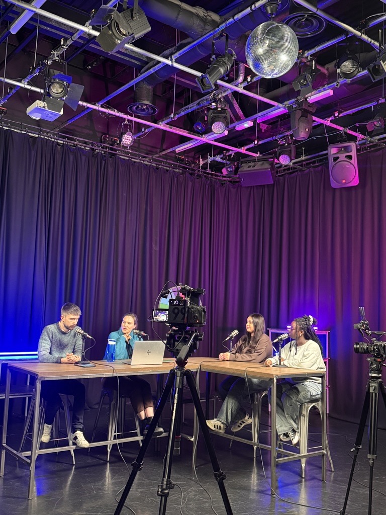 Four people seated at a desk during a livestream, smiling at the camera with laptops and notes in front of them. 
