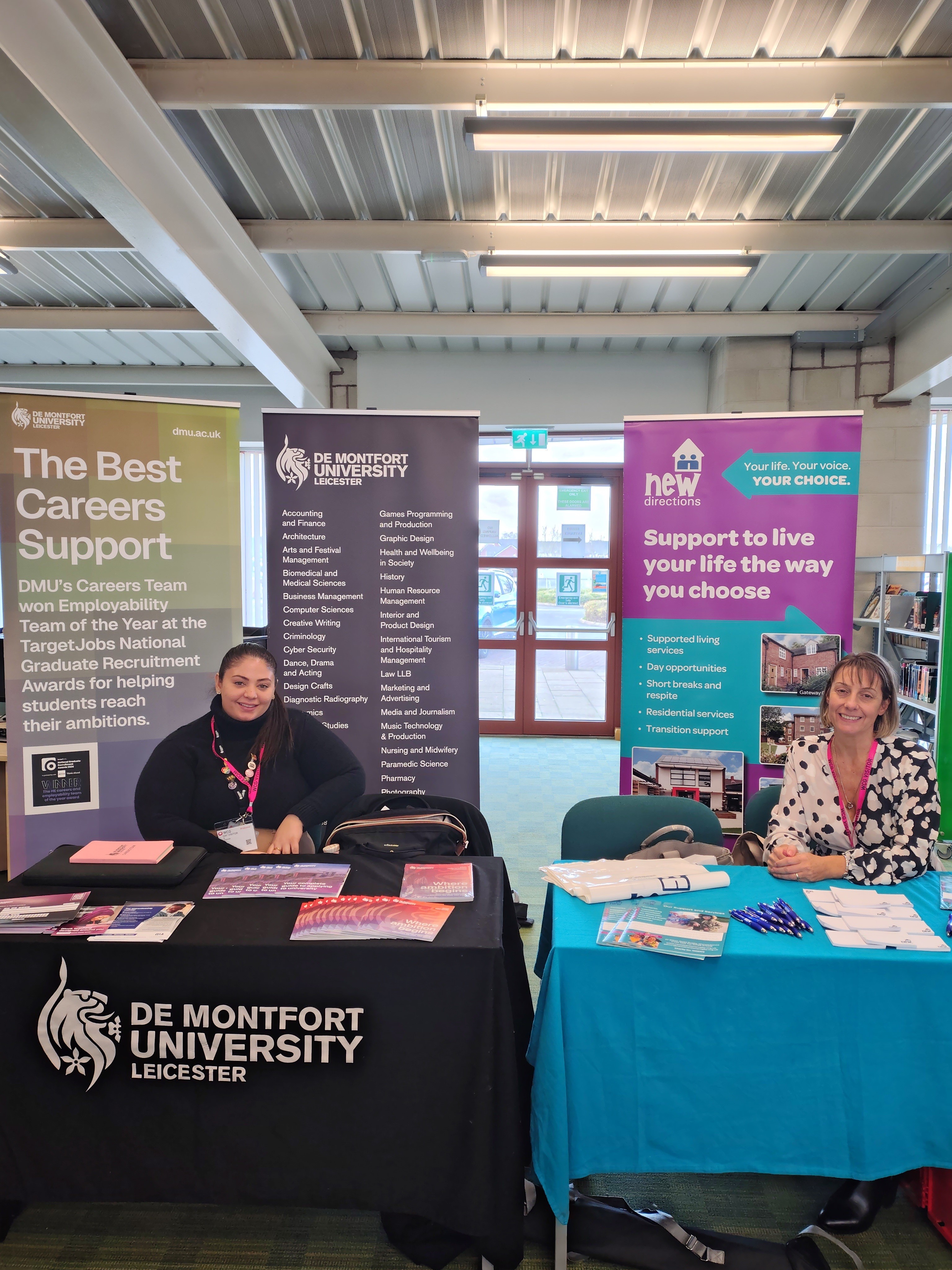 Evridiki from DMU smiling at the stand with course materials displayed on the table.