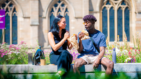 Male and female student sat eating outside Leicester Cathedral